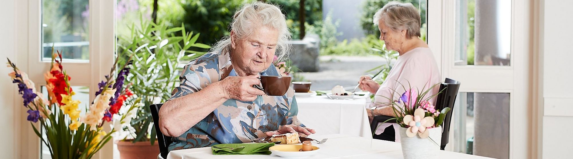 Eine Bewohnerin trinkt Kaffee am Frühstückstisch, der Blick geht durch die geöffnete Türe in den Garten mit viel Grün und schönen Pflanzen.