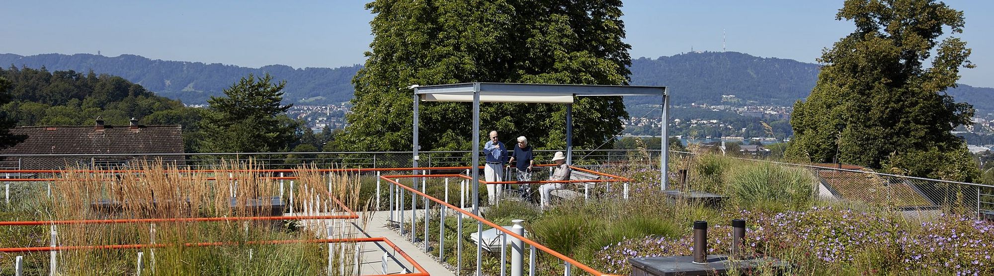 Blick auf den Garten der Mühlehalde mit Gehwegen und orangefarbenen Handläufen, im Pavillon sind drei Bewohnerin in Gespräch, der Blick vom Garten geht über die Dächer von Zürich.