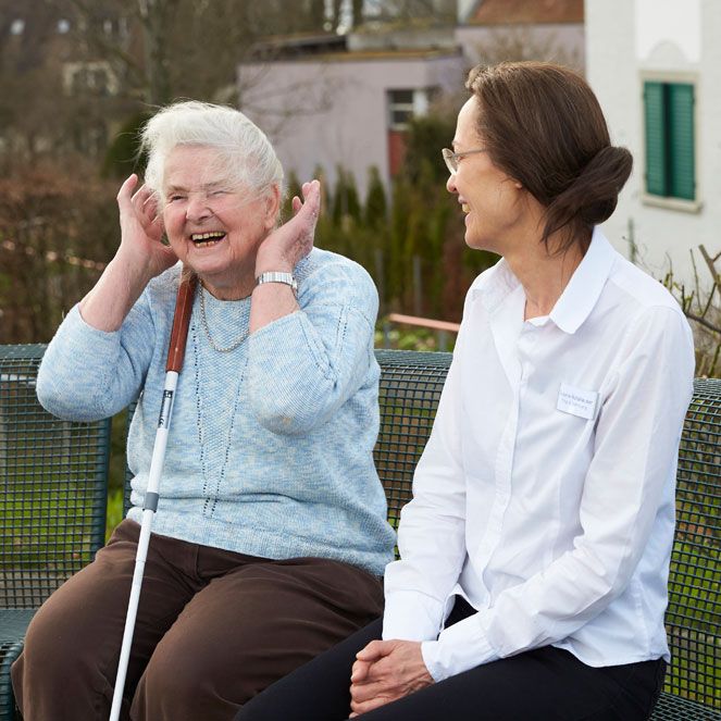 Eine lachende Bewohnerin mit Langstock sitzt im Garten der Mühlehalde, im Gespräch mit einer Pflegefachfrau, ebenfalls lachend.